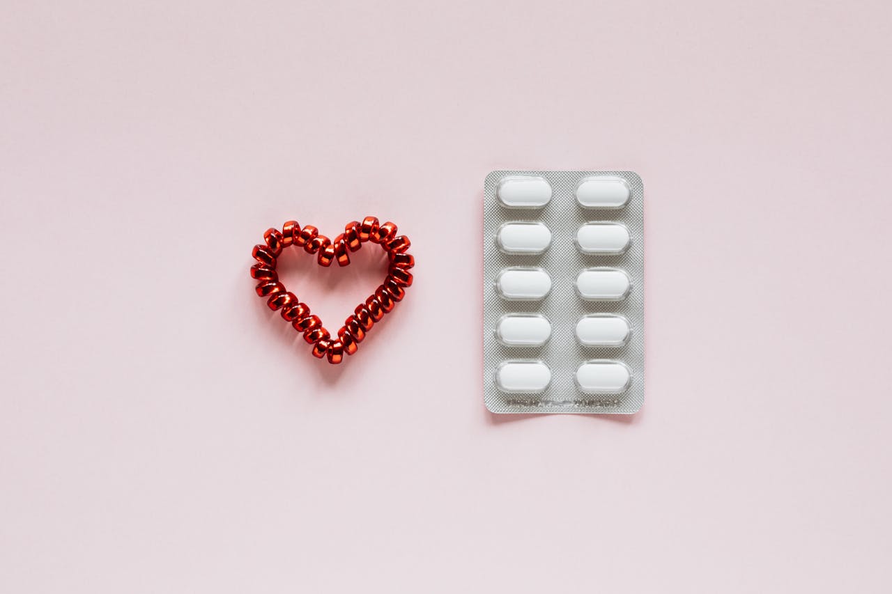 Flat lay of white pills and red beads forming a heart shape on a pink background.